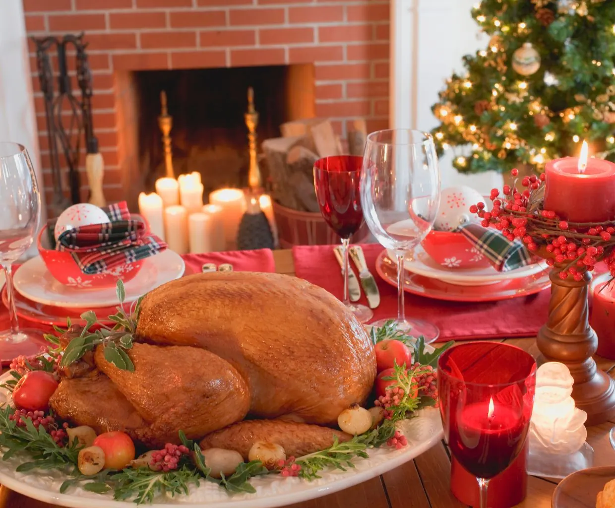 Mesa de cena de Navidad con pavo asado, guarniciones y velas rojas, con chimenea encendida y árbol decorado al fondo