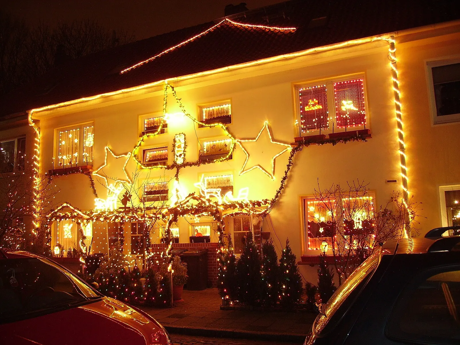 Fachada de una casa cubierta de luces navideñas cálidas, con estrellas luminosas y abetos iluminados en el jardín