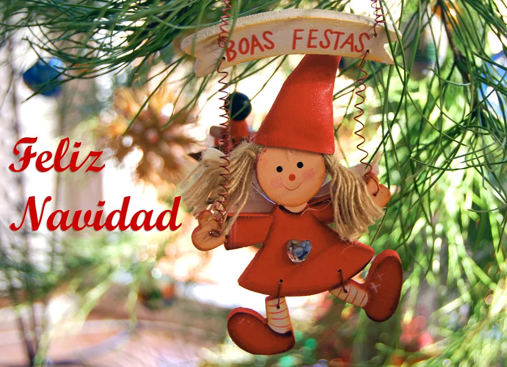 Muñeco de elfo con gorro rojo junto a carteles de «Feliz Navidad» y «Boas Festas» bajo un árbol decorado