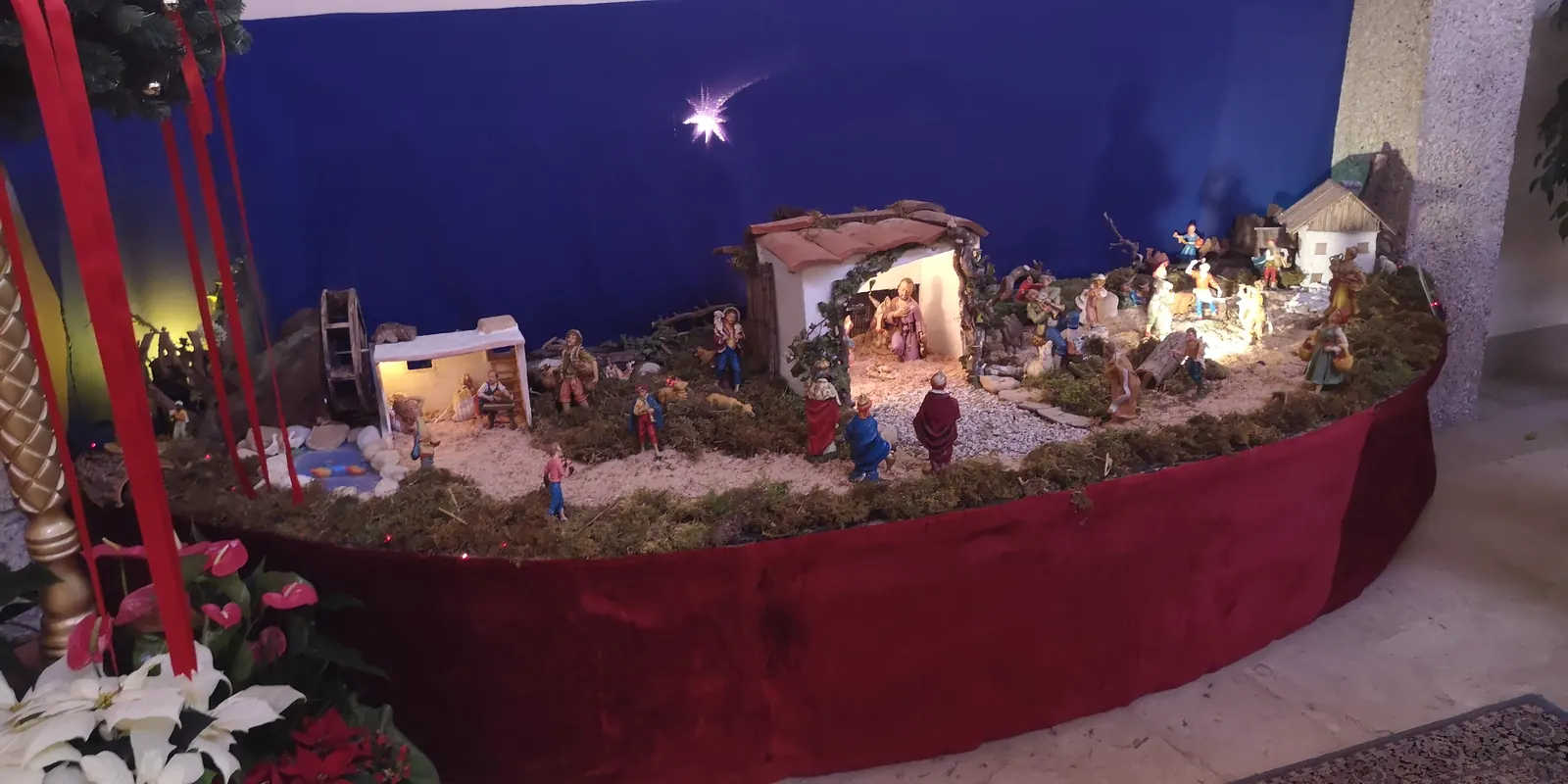 Belén con portal iluminado, figuras de pastores y Reyes, casitas, molino, río y musgo sobre una base de terciopelo rojo