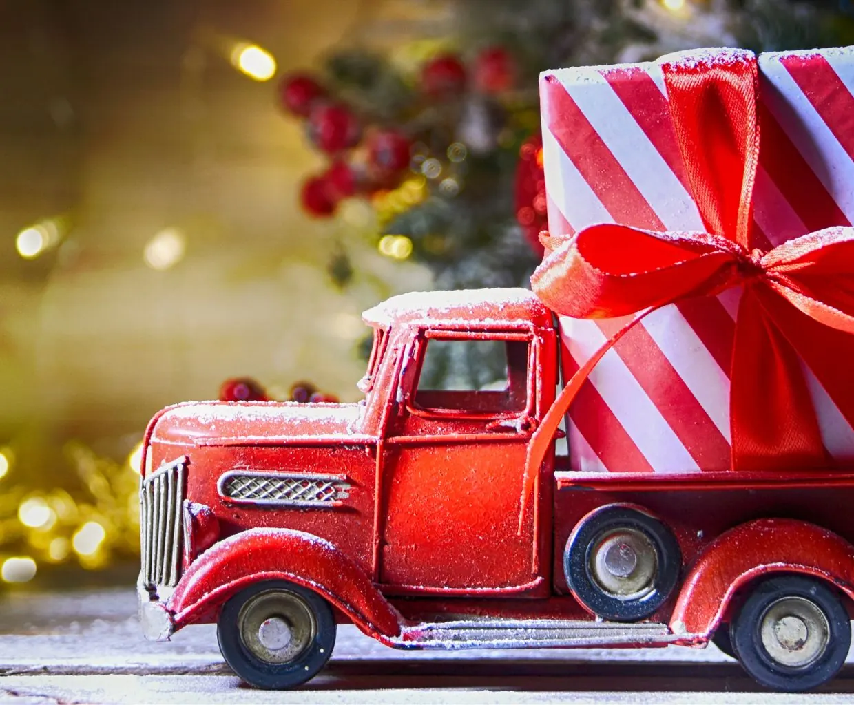 Camioneta roja de juguete estilo vintage cargada de regalos de Navidad envueltos, con luces cálidas desenfocadas detrás