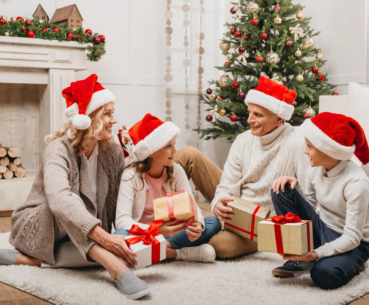Familia con gorros de Papá Noel sentada junto al árbol de Navidad intercambiando regalos