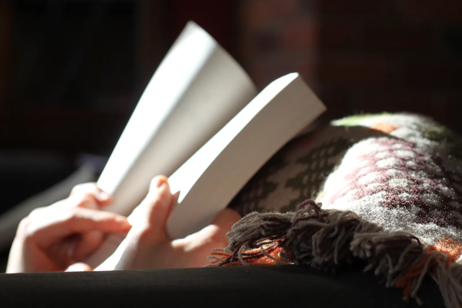 Manos sujetando un libro abierto junto a una manta de lana con flecos, iluminados por la luz de una ventana