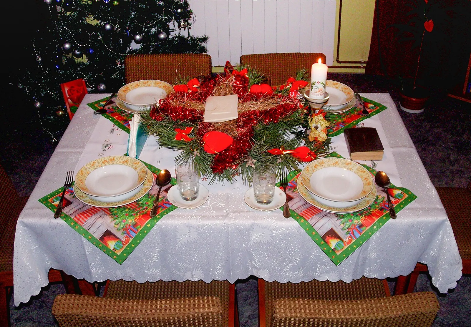 Mesa de Nochebuena con mantel blanco, centro de pino con lazos rojos, vela encendida y árbol de Navidad