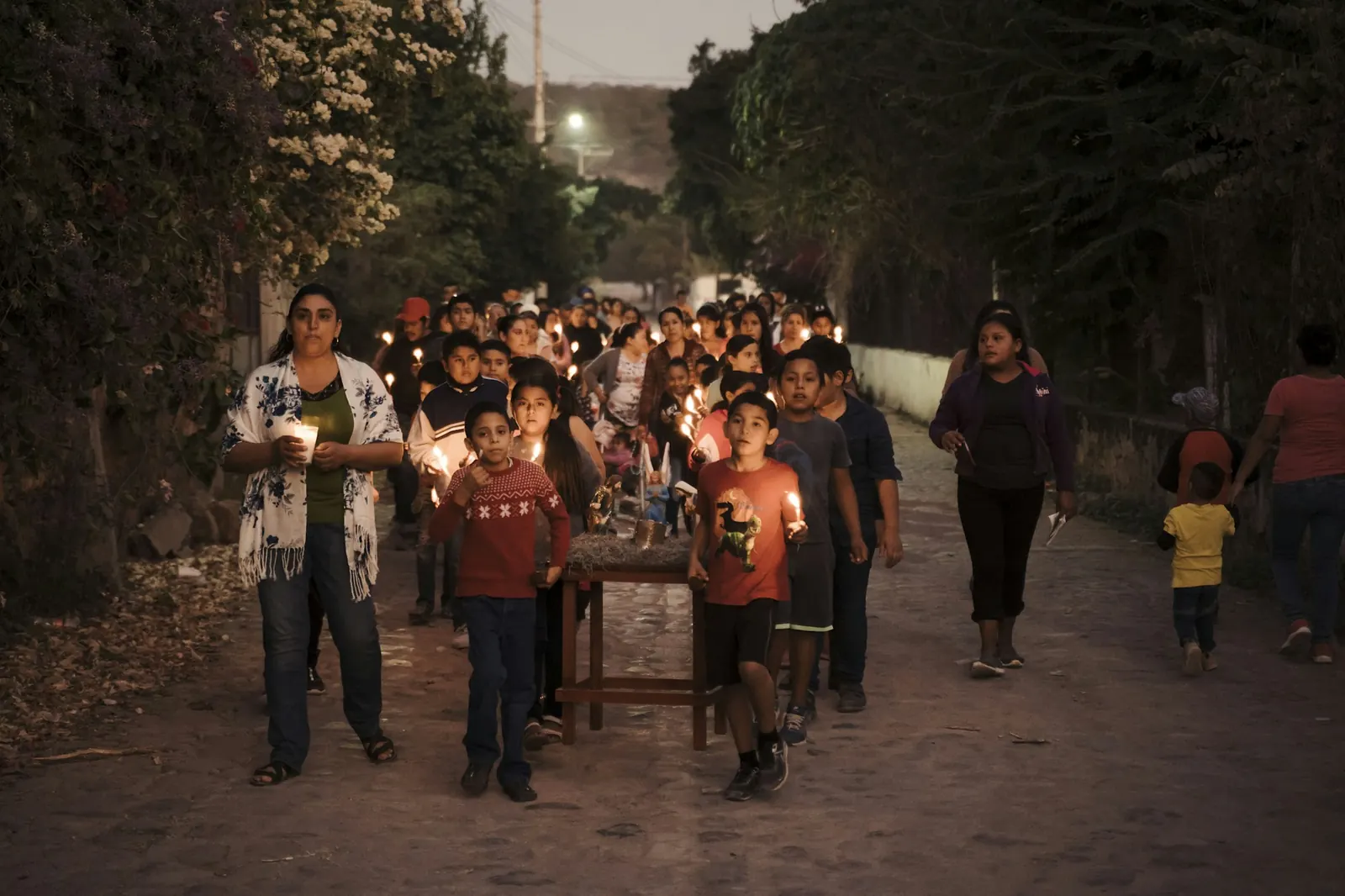Niños y adultos con velas encendidas recorren una calle al anochecer llevando en una mesita las figuras de María y José