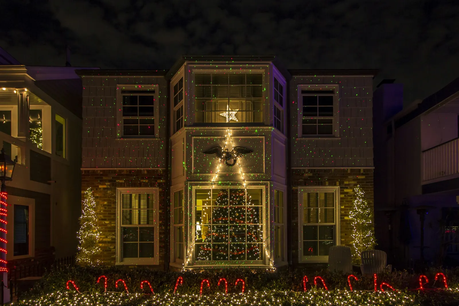 Fachada de una casa de noche cubierta de puntos de luz láser rojos y verdes, con un árbol iluminado en la ventana