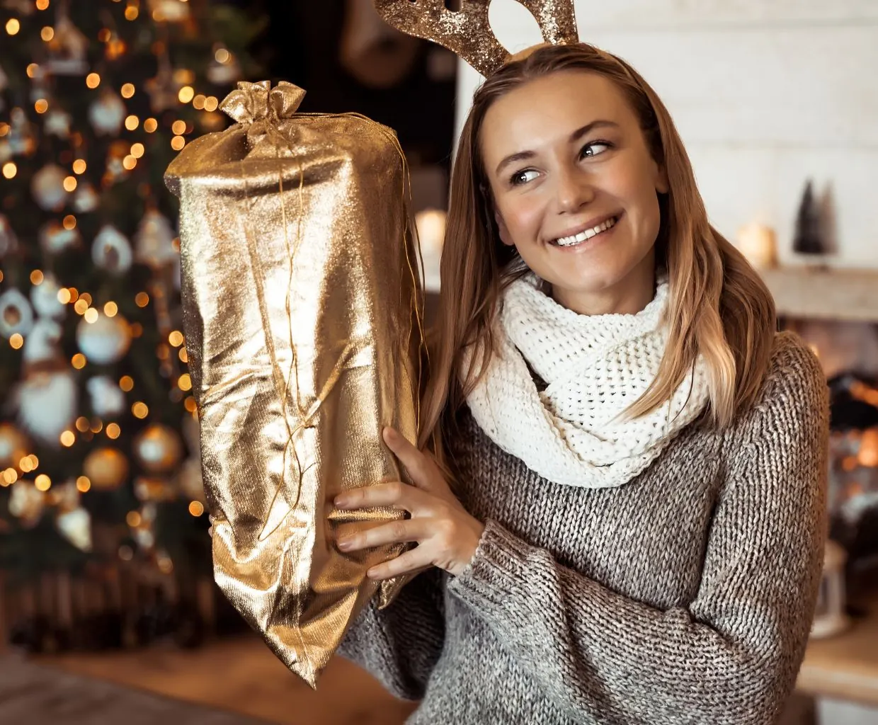 Mujer sonriente con bufanda color crema sostiene un gran regalo envuelto en dorado delante de un árbol de Navidad decorado