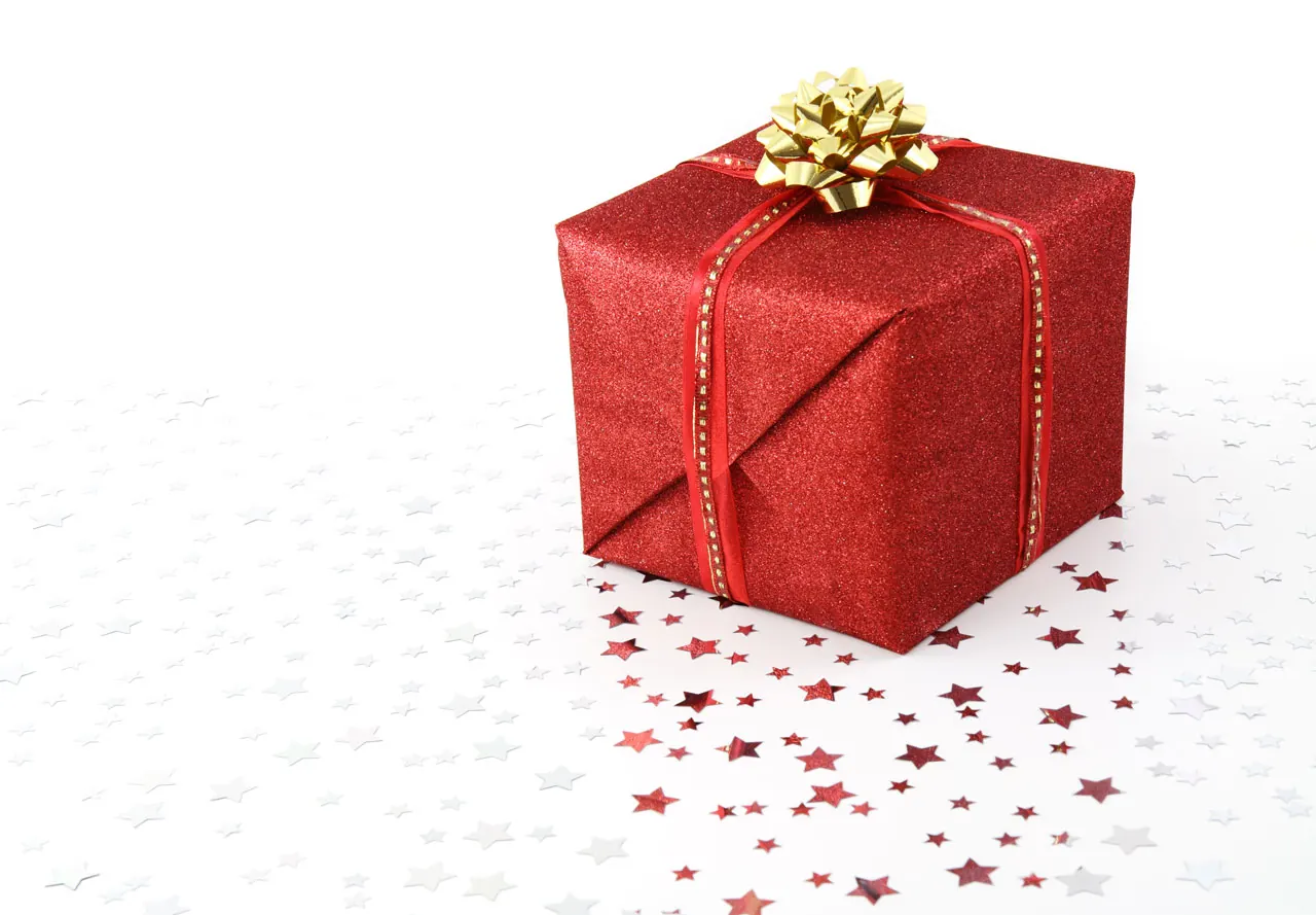 Caja de regalo envuelta en papel rojo brillante con cinta y lazo dorado, sobre fondo blanco con confeti de estrellas