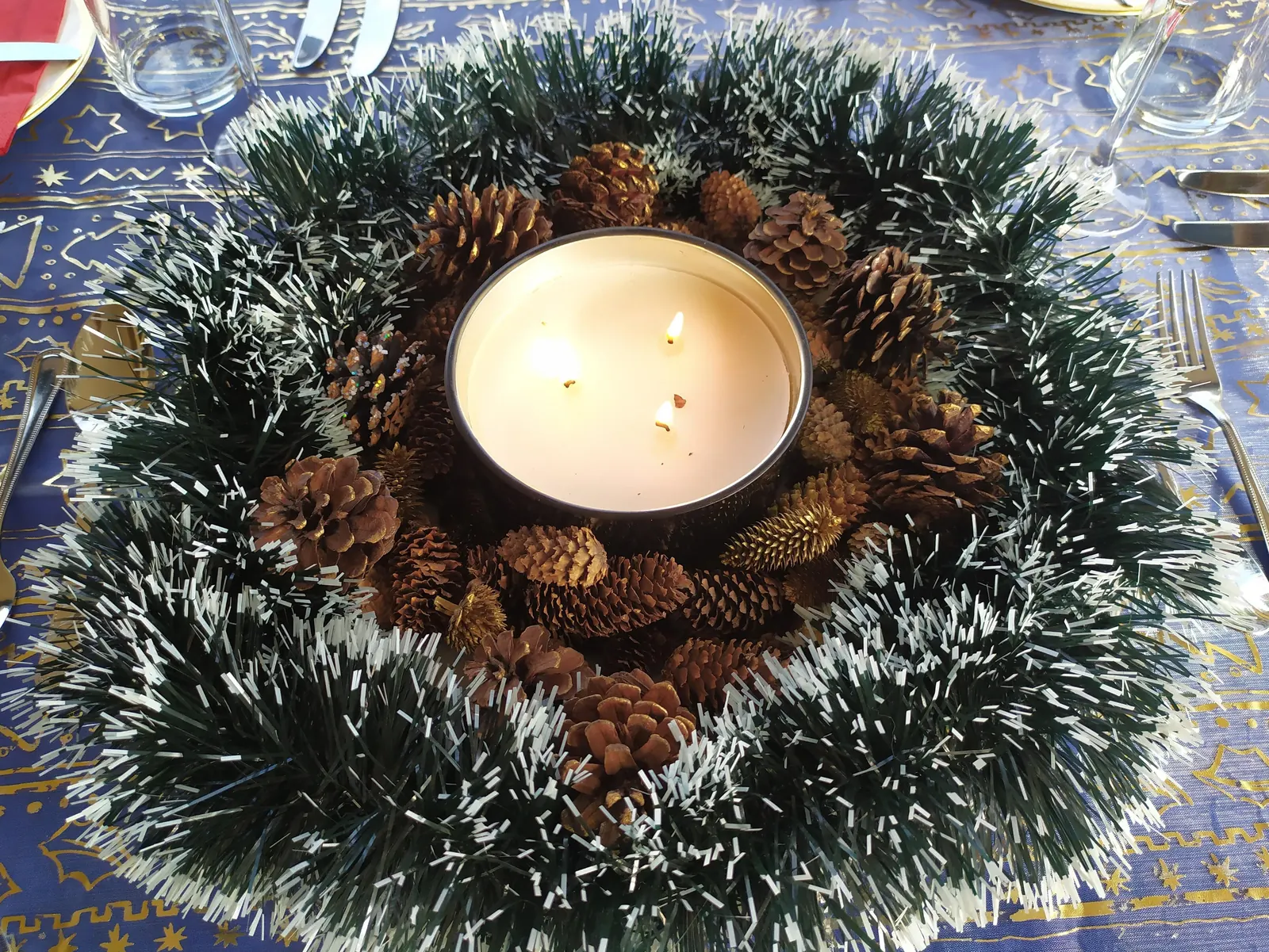 Centro de mesa navideño con una vela de tres mechas encendida rodeada de piñas y espumillón verde