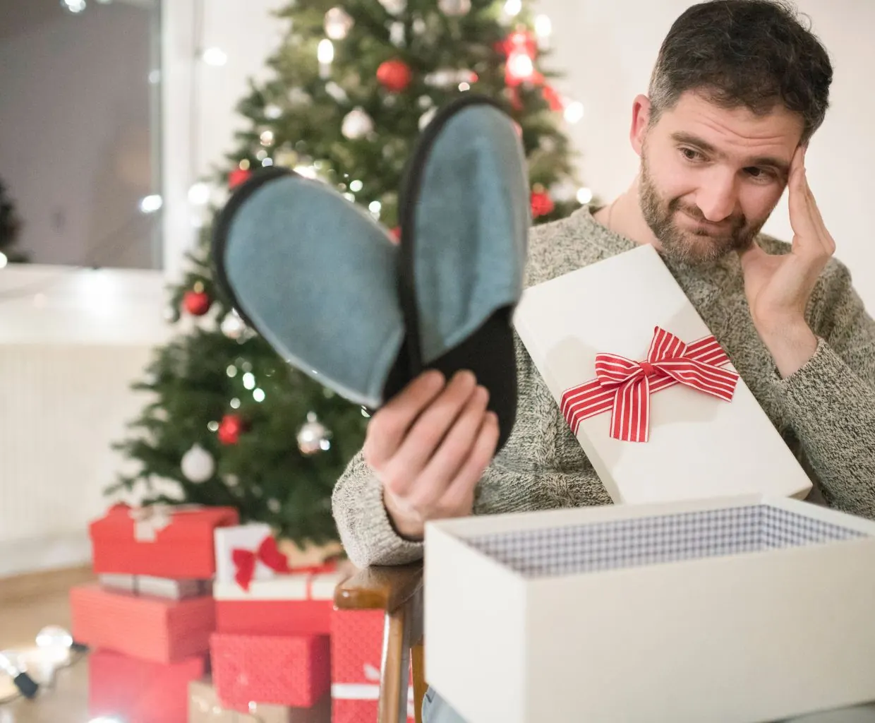 Hombre con barba saca unas zapatillas de casa de una caja de regalo con lazo rojo junto al árbol de Navidad