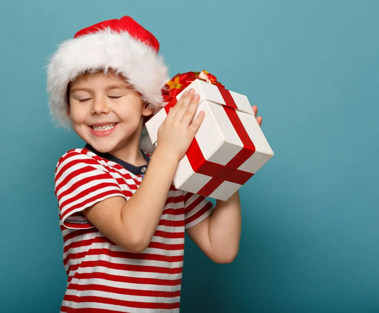 Niño sonriente con gorro de Papá Noel y jersey de rayas rojas y blancas abrazando un regalo envuelto