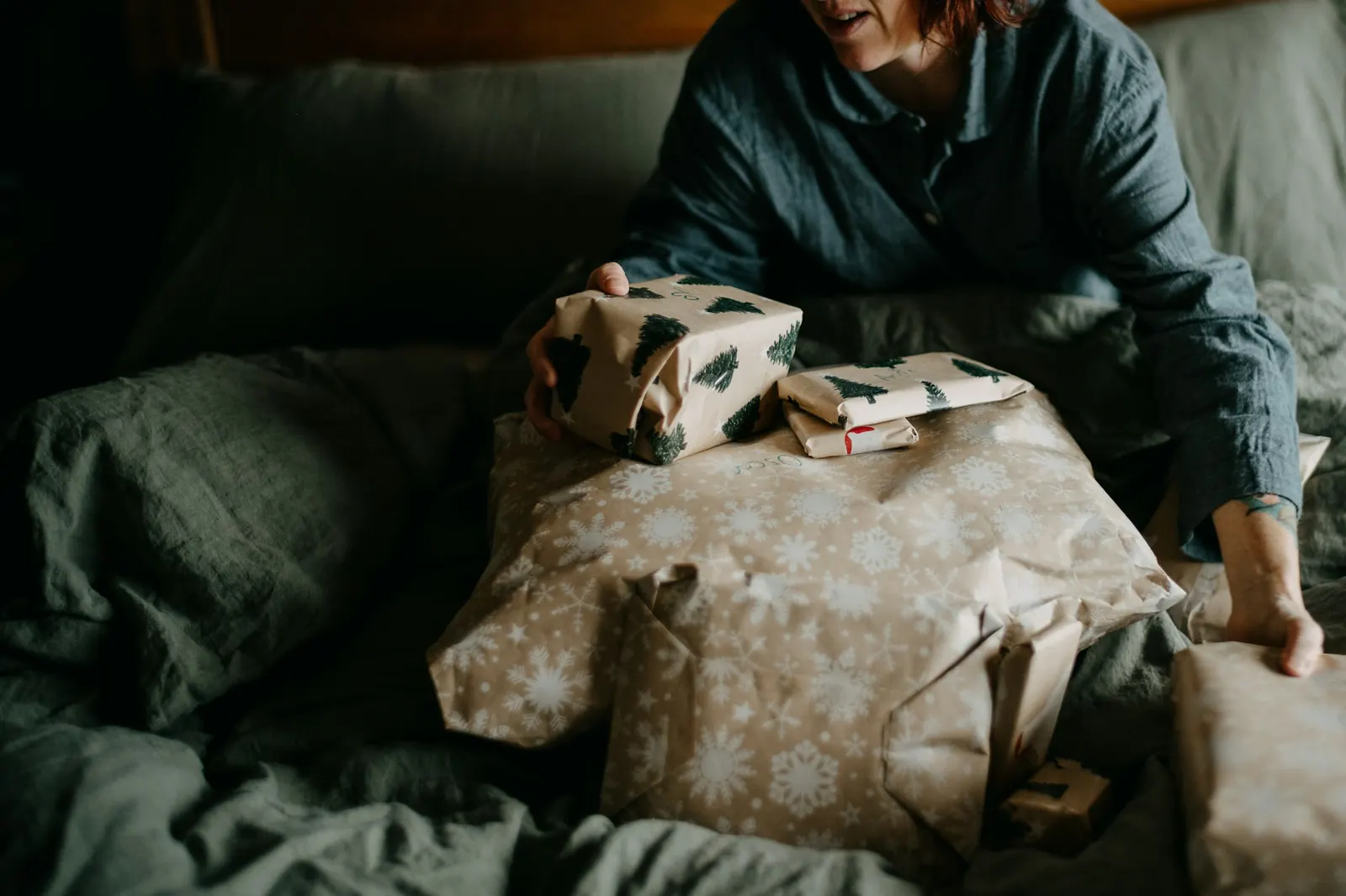 Mujer sentada en la cama entre sábanas verdes sostiene regalos envueltos en papel kraft con árboles y copos de nieve