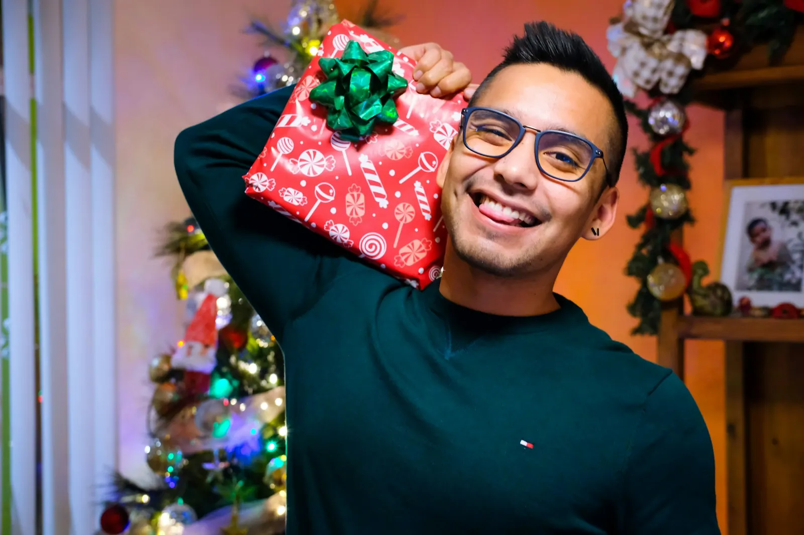 Chico sonriente con gafas sostiene sobre el hombro un regalo rojo con lazo verde delante de un árbol de Navidad