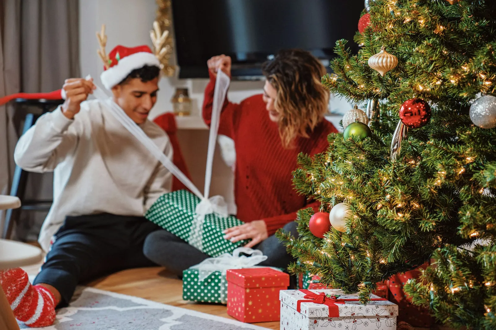 Una pareja sentada junto al árbol de Navidad abre un regalo de papel verde de lunares; él lleva gorro de Papá Noel