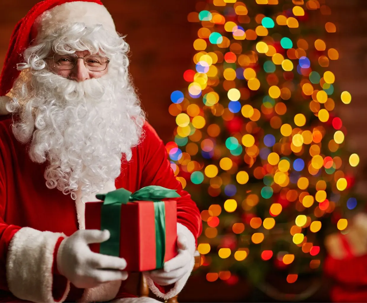 Papá Noel sosteniendo un regalo envuelto con lazo frente a un árbol con luces