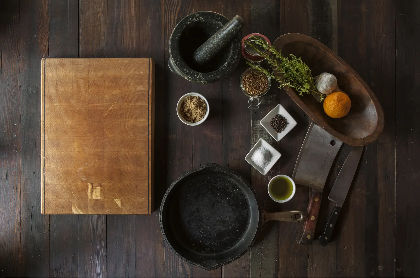 Tabla de cortar de madera, sartén de hierro, mortero, cuchillos, especias, romero y ajo sobre una mesa oscura