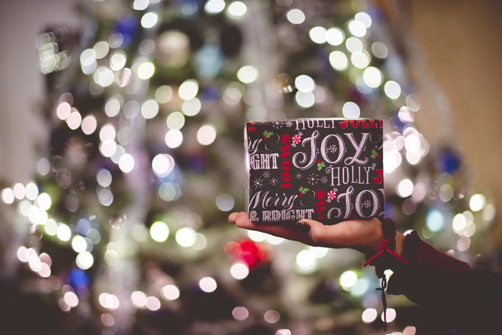 Mano que sostiene un pequeño regalo envuelto en papel navideño delante de un árbol con luces desenfocadas