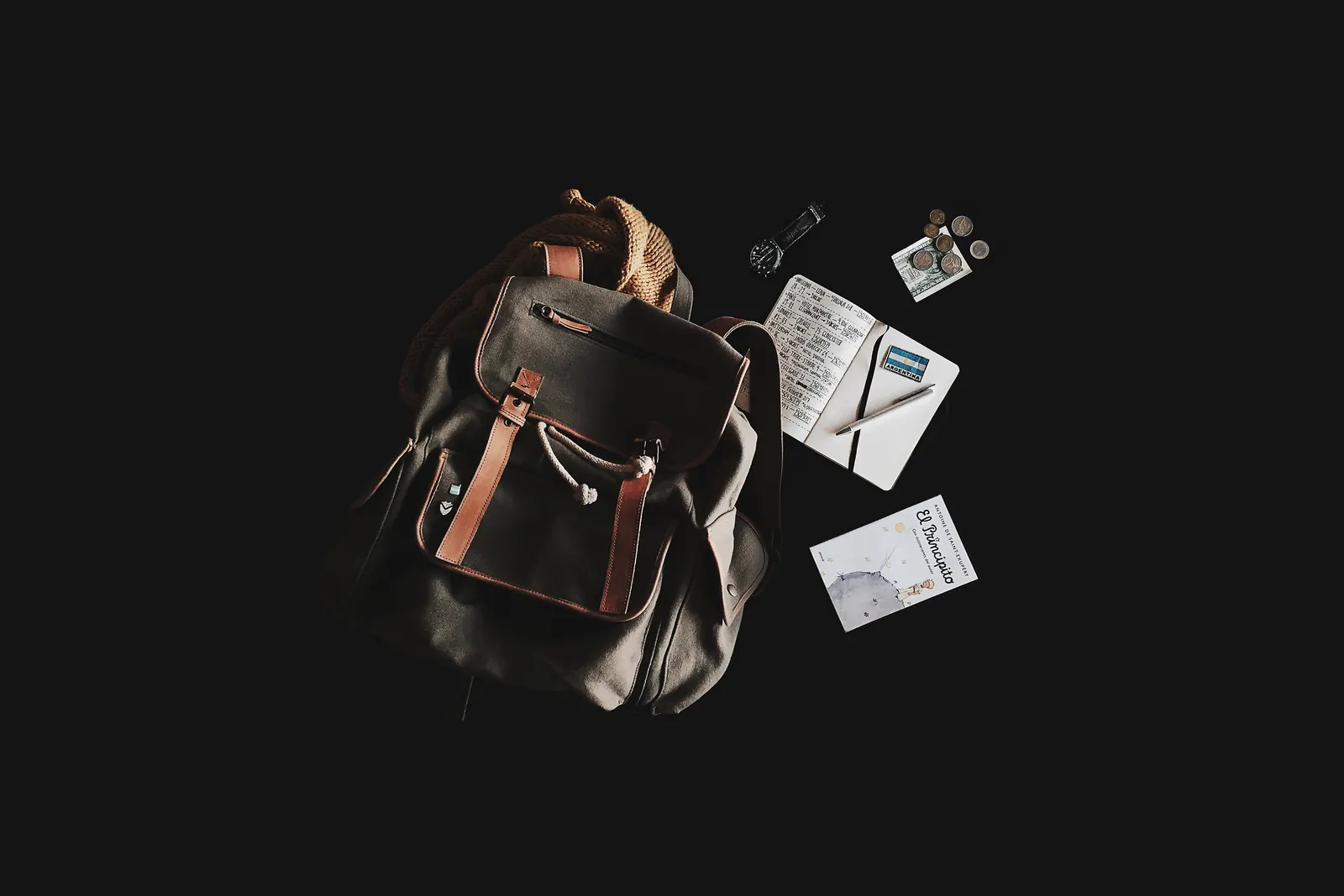 Mochila de lona con correas de cuero junto a un cuaderno de viaje, un reloj, monedas y el libro El Principito