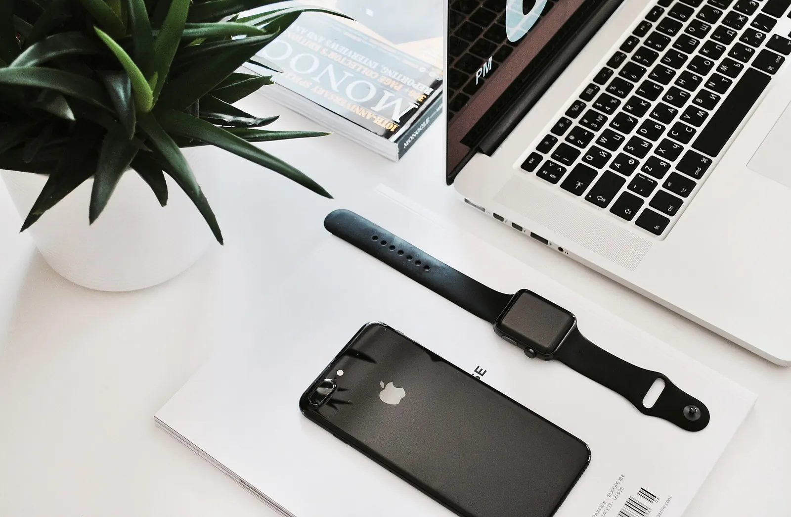 Mesa blanca con un portátil abierto, un reloj inteligente, un teléfono móvil y una planta en maceta