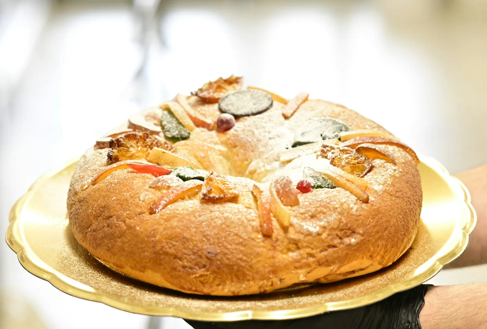 Roscón de Reyes con fruta confitada y azúcar glas sobre una bandeja dorada sostenida con guantes negros