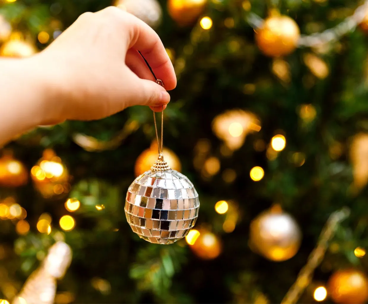 Mano colgando una bola de espejo tipo discoteca en un árbol de Navidad iluminado con luces cálidas desenfocadas