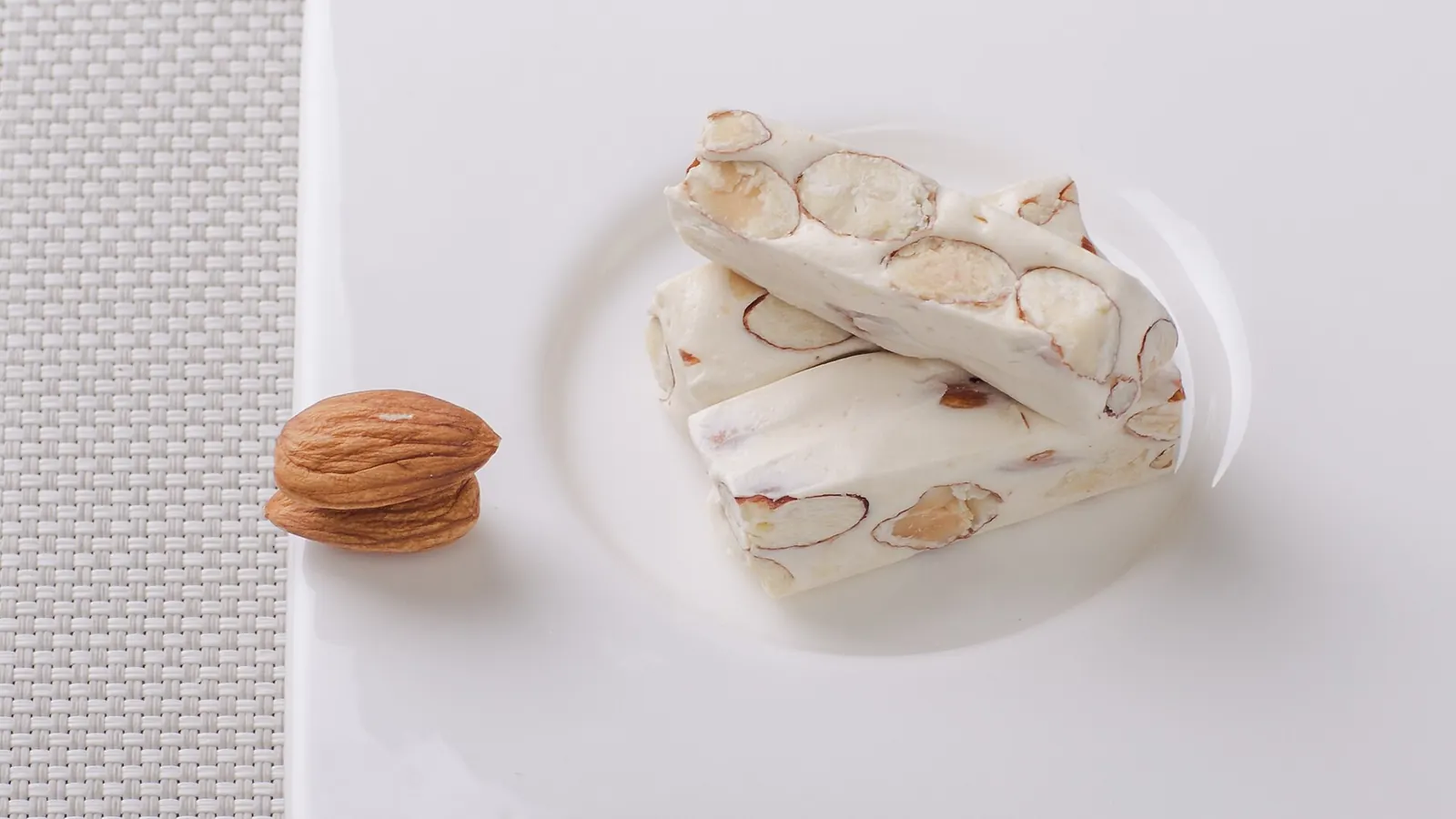Barritas de turrón blanco con almendras enteras en un plato blanco, junto a una almendra